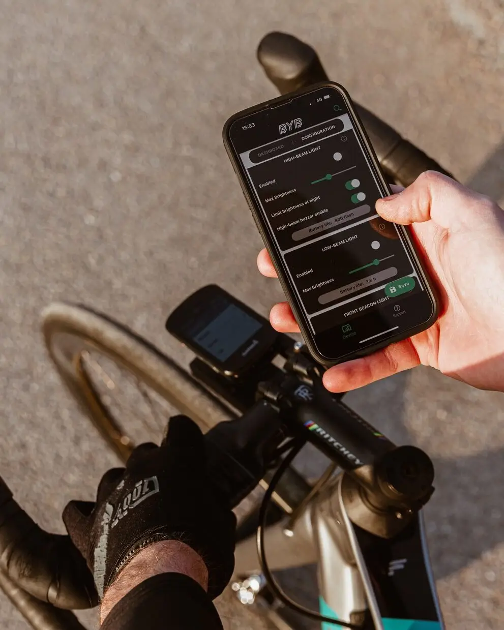 A rider holding a phone above the handlebar with the BYB app open on its configuration screen, showing the high-beam and low-beam sections with brightness sliders, a night-dimming toggle and a battery-life readout.